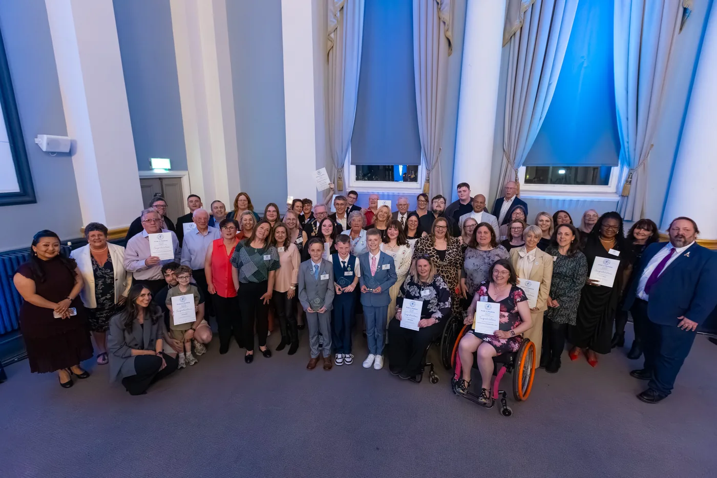 A large group of people holding certificates and trophies, photographed together in a hall with tall windows and blue curtains