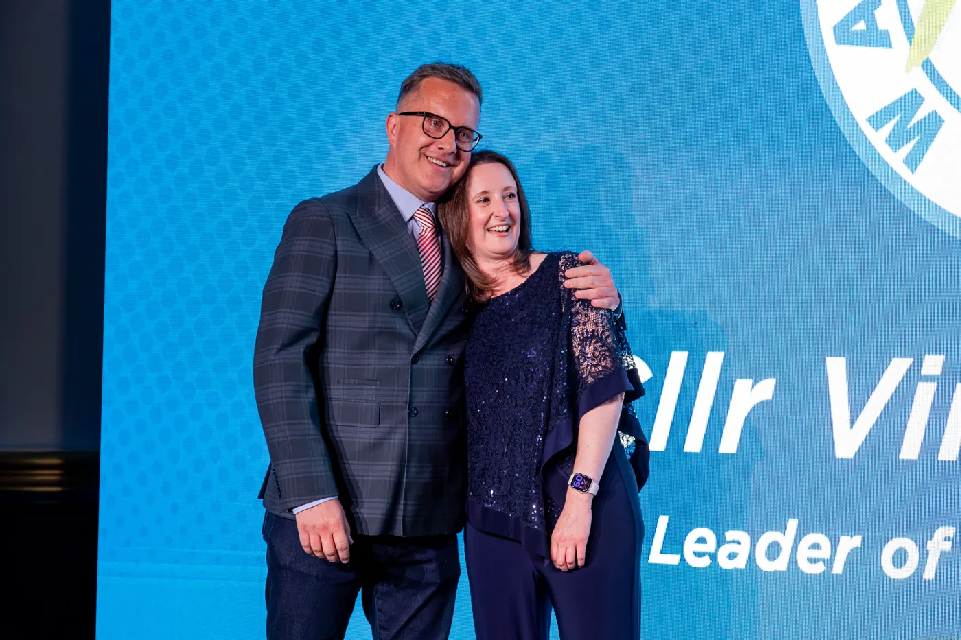 Two people standing together on a stage, smiling, in front of a blue screen showing the words Cllr Vin and Leader of